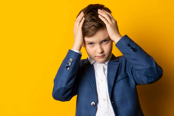 Ένα μικρό αγόρι με μπλε κοστούμι και λευκό πουκάμισο, όμορφα ντυμένο, βάζει τα χέρια στο κεφάλι του σε απογοήτευση ή απελπισία. Απομονωμένα σε κίτρινο φόντο. - Φωτογραφία, εικόνα