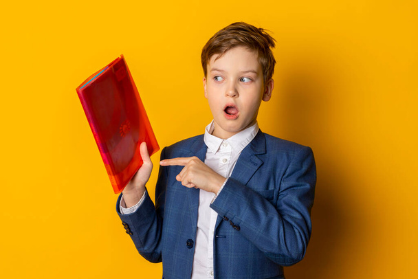 Ein überraschter kleiner Junge in blauem Anzug und weißem Hemd, ordentlich gekleidet, zeigt in der anderen Hand auf sein Lehrbuch. Vereinzelt auf gelbem Hintergrund. - Foto, Bild