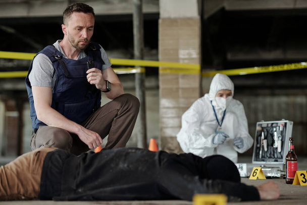 Молодой полицейский в пуленепробиваемом жилете, сидящий рядом с трупом, осматривая место преступления в окружении желтой ленты. - Фото, изображение