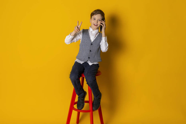 Ein fröhlicher kleiner Junge in grauer Weste und weißem Hemd, ordentlich gekleidet, telefonierend und ein Stück zeigend. Vereinzelt auf gelbem Hintergrund. - Foto, Bild