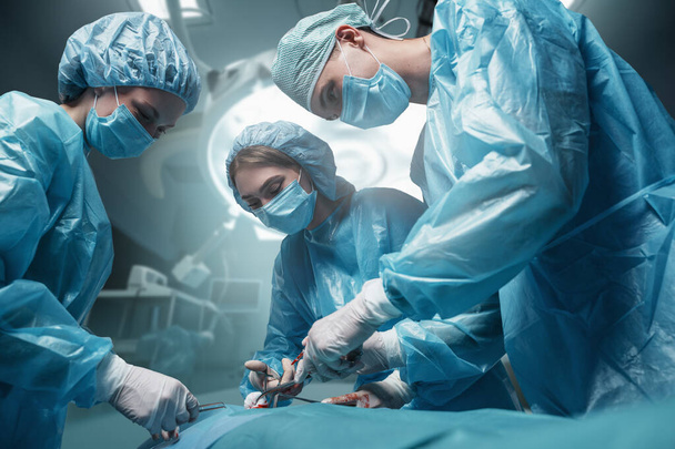 Tiro de tres cirujanos vestidos con túnicas y máscaras con paciente en quirófano. - Foto, imagen