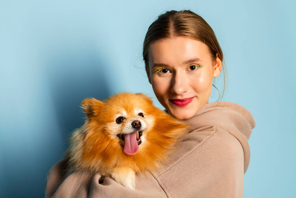 Çekici, gülümseyen genç bir kız ya sevimli Pomeranya köpeğini kucaklıyor ya da dilini dışarı çıkartıyor. Mavi arkaplanda izole. - Fotoğraf, Görsel