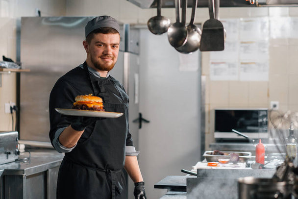 Otel restoranında profesyonel bir mutfak. Tatmin olmuş bir aşçı tabakta yeni hazırlanmış bir burger gösteriyor. - Fotoğraf, Görsel