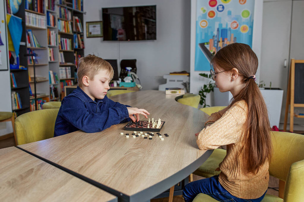 Biały chłopiec i dziewczyna grający w szachy, siedzący razem przy stole w szkole. Małe przyjacielskie koleżanki. - Zdjęcie, obraz