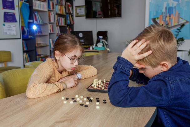 Niño y niña caucásicos jugando al ajedrez, sentados juntos en la mesa de la escuela. Pequeños compañeros de clase. - Foto, imagen