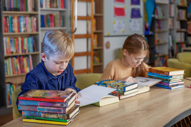 Μικρά παιδιά του σχολείου διαβάζουν βιβλία μαζί, ενώ κάθονται στο τραπέζι στη βιβλιοθήκη. Αγόρι και κορίτσι σπουδάζουν στο σχολείο. Παγκόσμια Ημέρα Βιβλίου - Φωτογραφία, εικόνα