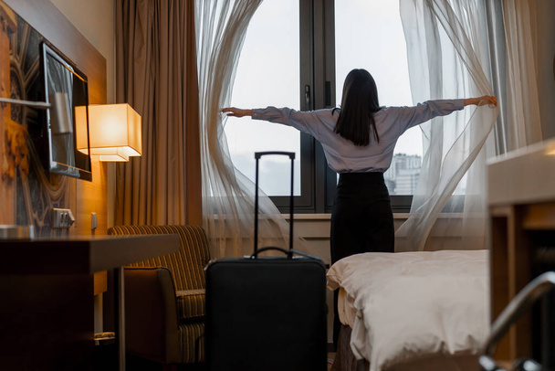 young woman opening curtain while standing luxury hotel room looking through window enjoying city skyscrapers close up - Photo, image