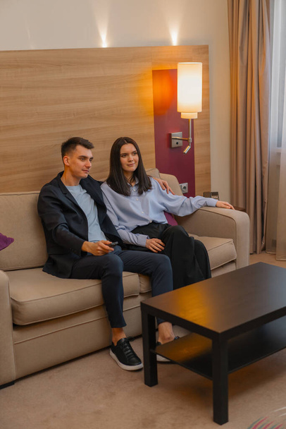 joven pareja habitación de hotel sala de estar viendo la televisión juntos El hombre toma el control remoto y cambia los canales - Foto, Imagen