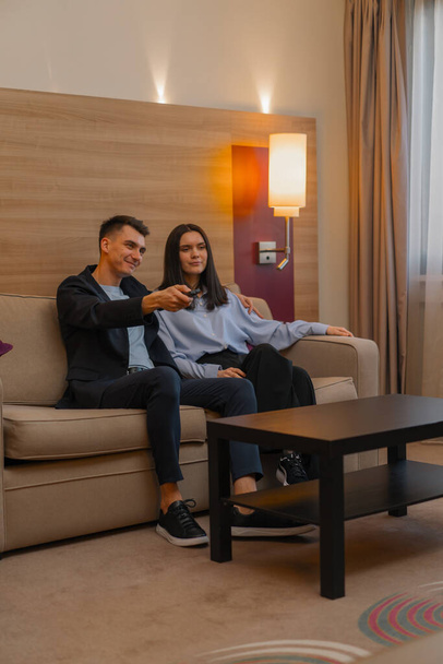 Nuori pari hotellihuone olohuone katsomassa televisiota yhdessä Mies ottaa kaukosäätimen ja vaihtaa kanavia - Valokuva, kuva