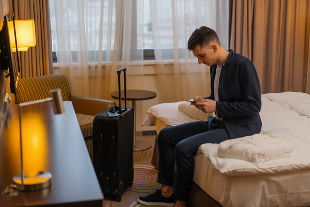 Man with suitcase sitting on bed in hotel room while traveling recreation concept - Foto, Imagen