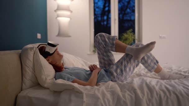 Příjemně se usmívající žena leží na posteli v pyžamu s virtuální realitou sluchátka ponořen do metaverzní přehrávání videohry sledovat film vychutnat čas doma. Žena ve 3D brýlích si pro zábavu vybere VR menu. - Záběry, video