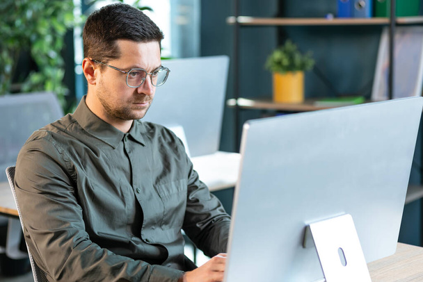 Hombre barbudo guapo serio en camisa casual sentado en el lugar de trabajo y trabajando con la computadora - Foto, imagen