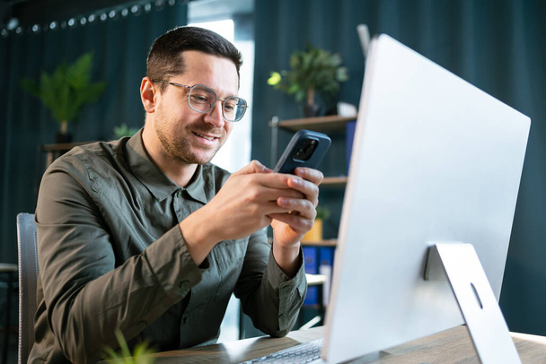 Foto de hombre adulto sonriente de 30 años en camisa verde usando computadora portátil y teléfono móvil, mientras está sentado en la mesa y trabajando en la oficina - Foto, imagen