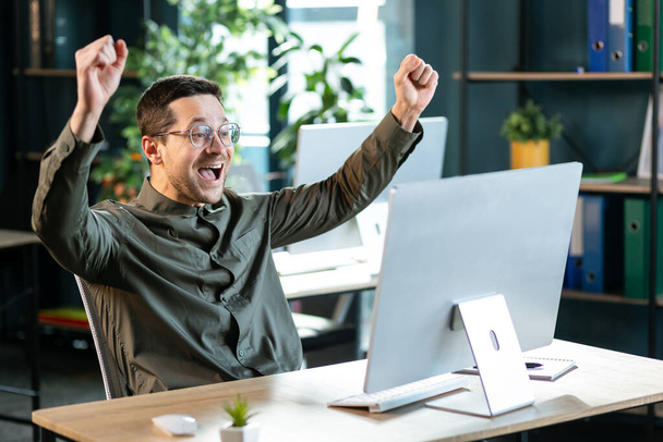 Young guy making YES gesture in front of computer at office. Millennial man reaching success, working with portable pc, making profitable deal, signing online contract, celebrating achievement - Фото, изображение