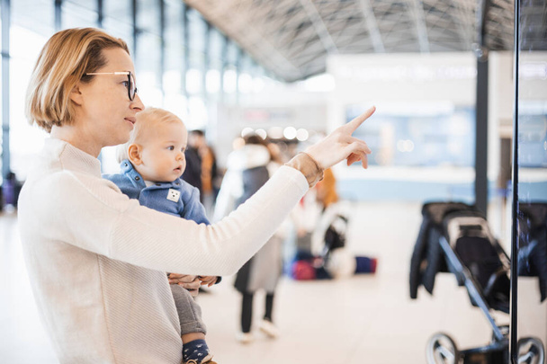 Μητέρα που ταξιδεύει με παιδί, κρατάει το αγοράκι της στο αεροδρόμιο, ελέγχει το πρόγραμμα πτήσεων, περιμένει να επιβιβαστεί στο αεροπλάνο. Ταξίδι με τα παιδιά έννοια - Φωτογραφία, εικόνα