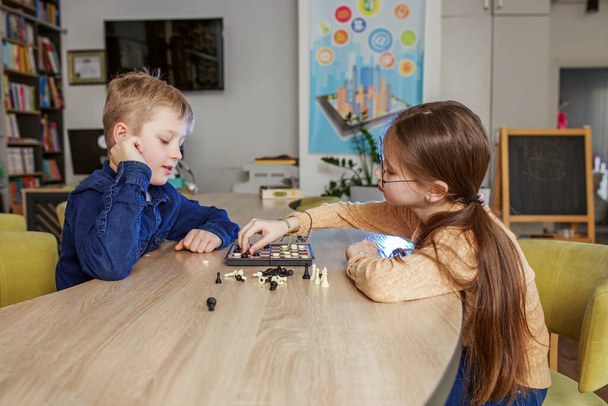 Kaukasische Jungen und Mädchen spielen Damesteine und sitzen zusammen an einem Tisch in der Schule. Wenig freundliche Klassenkameraden. - Foto, Bild