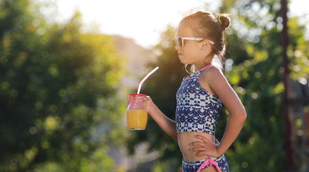 Beautiful little girl, cute toddler with sunglasses is drinking orange juice and shows thumb up. Kid in in swimwear having fun during family vacation in a resort near pool. summer holidays. - Foto, Imagem