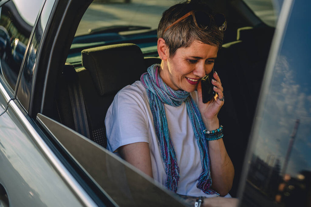 una mujer madura mujer caucásica sentada en el asiento trasero del coche que trabaja en el ordenador portátil hacer una llamada telefónica hablar en el día de verano con pelo gris corto moderno en la carretera con espacio de copia casual - Foto, imagen
