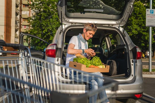 Jedna kobieta dojrzała kaukaska kobieta siedzi w bagażniku swojego samochodu na parkingu supermarketu lub sklepu spożywczego z warzywami w pudełku umieszczając je w pojeździe - Zdjęcie, obraz