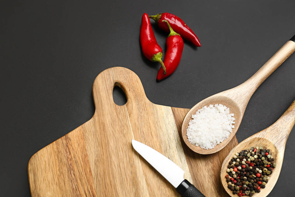 Wooden cutting board, spoons with spices and chili peppers on dark background, closeup - Fotó, kép
