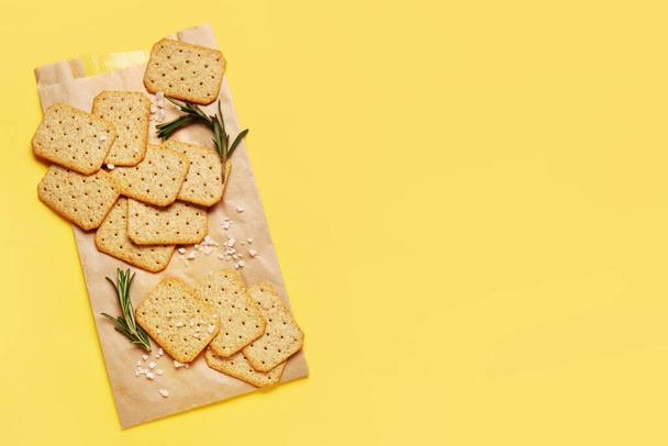 Parchment with tasty crackers, rosemary and salt on yellow background - Photo, Image