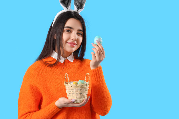 Young woman in bunny ears with basket of Easter eggs on blue background - Фото, изображение
