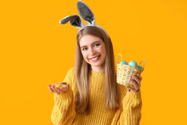 Young woman in bunny ears with basket of Easter eggs on yellow background - Foto, Bild