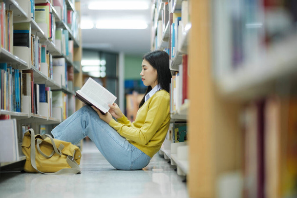 Νεαρή φοιτήτρια λυκείου με καθημερινά ρούχα κάθεται στο πάτωμα διαβάζοντας το βιβλίο, μελετώντας και κάνοντας έρευνα για το έργο του σχολείου σε μια βιβλιοθήκη. Μάθηση, Εκπαίδευση, Βιβλιοθήκη έννοια. - Φωτογραφία, εικόνα