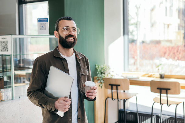 Hombre barbudo sonriente en vasos de pie sosteniendo portátil y taza de café en la cafetería mirando por la ventana. interior moderno - Foto, Imagen
