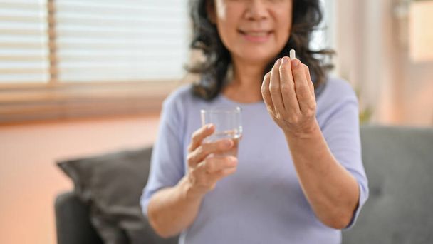 Imagem de close-up, uma mulher asiática aposentada de 60 anos de idade, segurando um copo de água e um comprimido, tomando um remédio em sua sala de estar. saúde, bem-estar, conceito de estilo de vida - Foto, Imagem