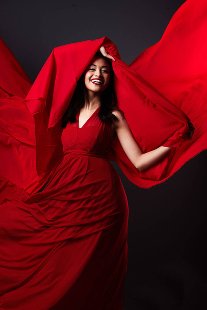 Woman, beauty and fashion, red art on dark background with energy and aesthetic movement. Flowing silk motion, fantasy and artistic model with smile in creative fabric designer dress in studio. - Foto, afbeelding