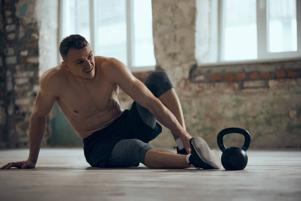 Feeling tired after hard training session. Young muscular man sitting on floor shirtless with sweating body. Concept of sportive lifestyle, body care, fitness, hobby, health, action and motion - Foto, afbeelding
