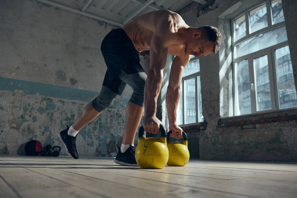 Young handsome man with muscular, strong, fit body training indoors with weights on a daytime. Daily training session. Concept of sportive lifestyle, body care, fitness, hobby, health - Photo, image