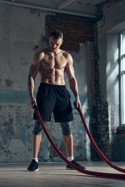 Full-length portrait of young muscular man with strong, relief, fit body posing with sport equipments indoors. Battle ropes for workout. Sportive lifestyle, body care, fitness, hobby, health concept - Фото, изображение