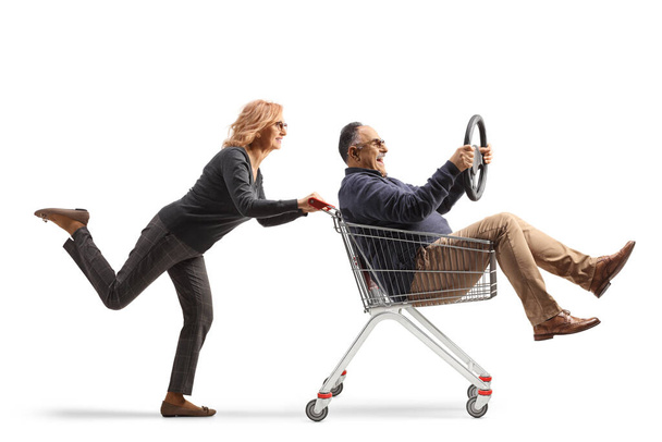 Woman pushing a mature man inside a shopping cart sitting and holding a steering wheel isolated on white background - Foto, immagini