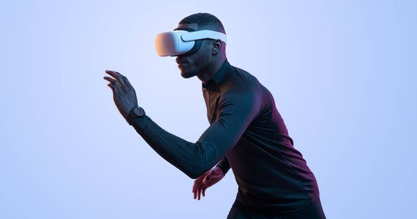 Side view of active young African American male athlete in black activewear running while exploring virtual reality in VR glasses against blue background - Photo, Image