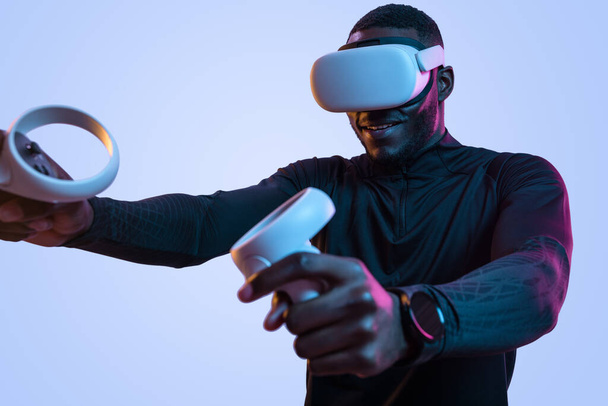 Positive bearded African American male gamer in black turtleneck and VR googles holding consoles while playing videogame near blue background - Photo, Image