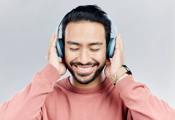 Happy, muziek en Aziatische man met koptelefoon, streaming en vrolijk op een grijze studio achtergrond. Japan, man en zelfverzekerde man met headset, luisteren naar audio en geluiden voor vrede, glimlach en wellness. - Foto, afbeelding