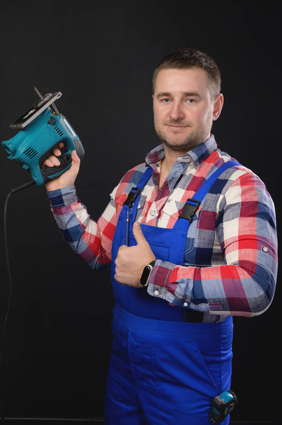Smiling man in a plaid shirt and overalls holds a jigsaw in his hand. on a black background and with the other hand shows a thumbs up gesture. - Foto, Bild