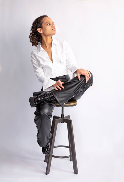 Modische Afrikanerin in weißem Hemd, Lederhose, hohen Stiefeln sitzend, posierend auf Stuhl. Studioporträt. - Foto, Bild