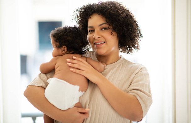 Happy loving young african american curly woman plus size holding baby in her arms in clinic or home interior. Mother and child, vaccination, child health care, motherhood and family - Photo, Image