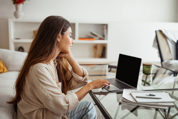 Smiling pretty millennial mixed race woman typing on laptop with empty screen in white living room interior. Work, business remotely, freelance at website and new normal at home - Photo, Image