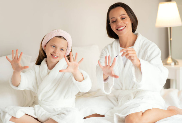 Glad young caucasian female, small girl in bathrobe sit on white soft bed, do manicure with nail polish, enjoy beauty care in bedroom interior. Weekend mom and daughter, relationship at home - Valokuva, kuva