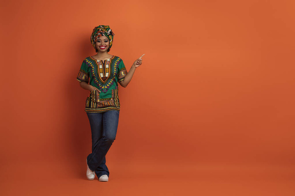 Positiv fröhlich schöne amerikanische Millennial-Amerikanerin in traditioneller Tracht mit einer Hand in der Tasche, die freien Platz für Werbung zeigt, orangefarbener Studiohintergrund, volle Länge - Foto, Bild