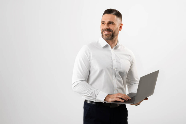 Successful middle aged man in formal wear using laptop over grey studio background, looking aside at free space. Positive businessman having online job, communicating via internet - Photo, Image