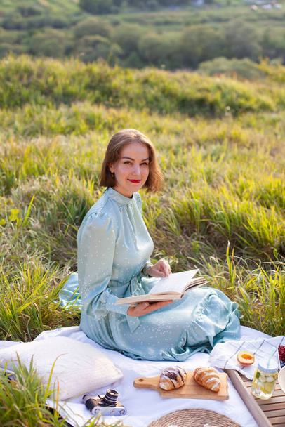 A woman in a long summer dress with short hair sitting on a white blanket with fruits and pastries and reading the book. Concept of having picnic in a city park during summer holidays or weekends.  - Foto, Imagen