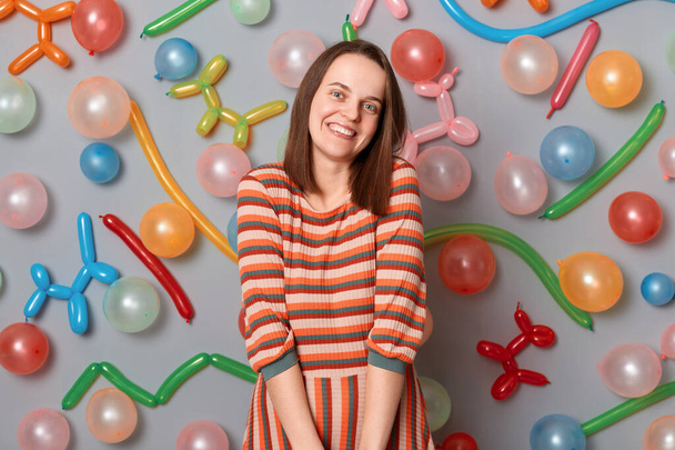 Portrait of cute smiling woman with brown hair wearing striped dress looking at camera with happy expression, being in festive mood, standing against gray wall with colorful balloons - Foto, Imagem