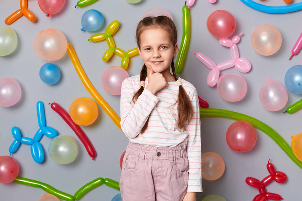 Image of cute charming smiling little girl with braids wearing casual clothing posing isolated over gray background with balloons, holding her chin, showing thumb up. - Foto, afbeelding