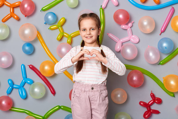 Portrait of lovely charming little girl with braids wearing casual clothing, showing heart shape with hands, posing isolated over gray background with balloons. - Foto, afbeelding