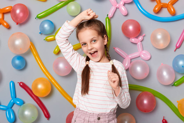 Photo of excited joyful little girl with braids wearing casual clothing clenched fists raised arms celebrating birthday dancing standing against gray wall with colorful balloons. - Photo, image